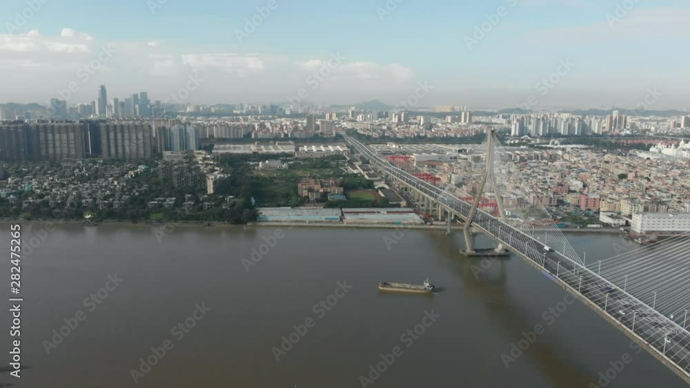 The drone is approaching the residential part of the city across the river. On the left side is a large bridge with heavy traffic. A large cargo ship sails along the river The drone is approaching the residential part of the city across the river. On the left side is a large bridge with heavy traffic. A large cargo ship sails along the river