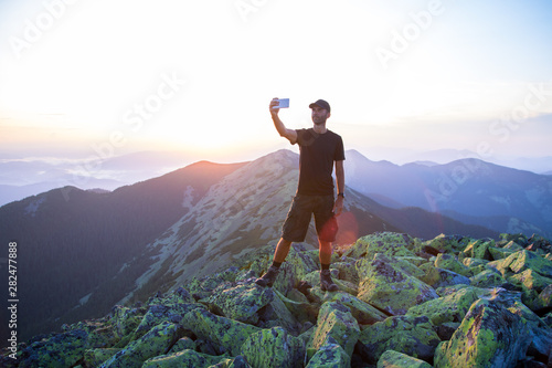 caucasian man hiker is on the peak of green stones mountain is making selfie photos on the sunset with a smartphone © AlexR