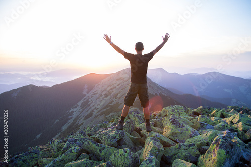 caucasian man hiker is on the peak of green stones mountain watching sunset with hands in the air