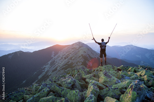 caucasian man hiker is on the peak of green stones mountain watching sunset with trekking sticks in the hands in the air