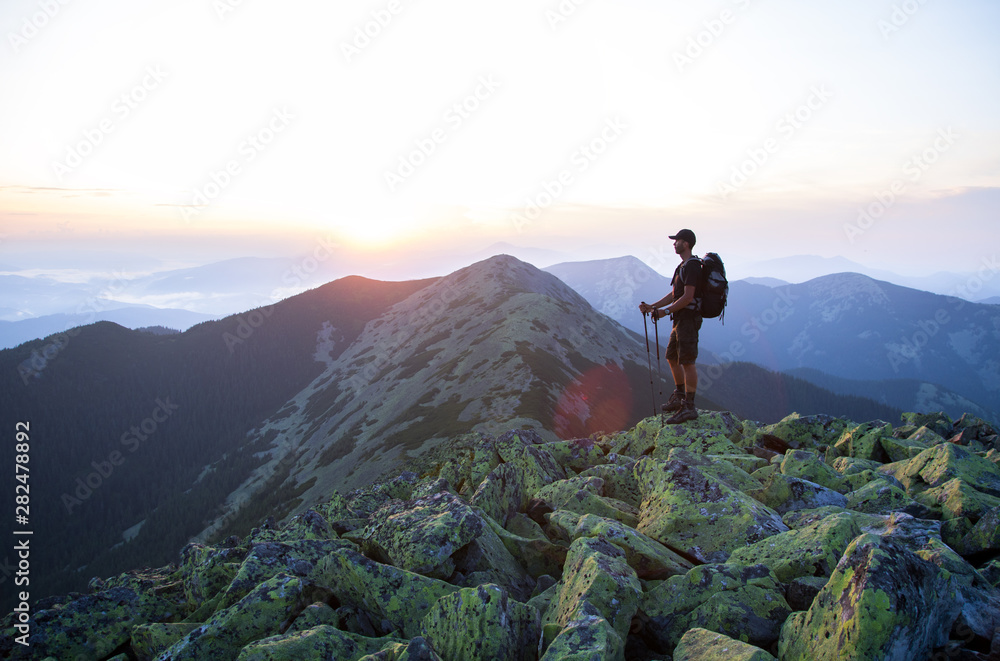 caucasian man hiker with backpack and trekking sticks is on the peak of green stones mountain enjoy a panoramic view of mountains in the sunset light