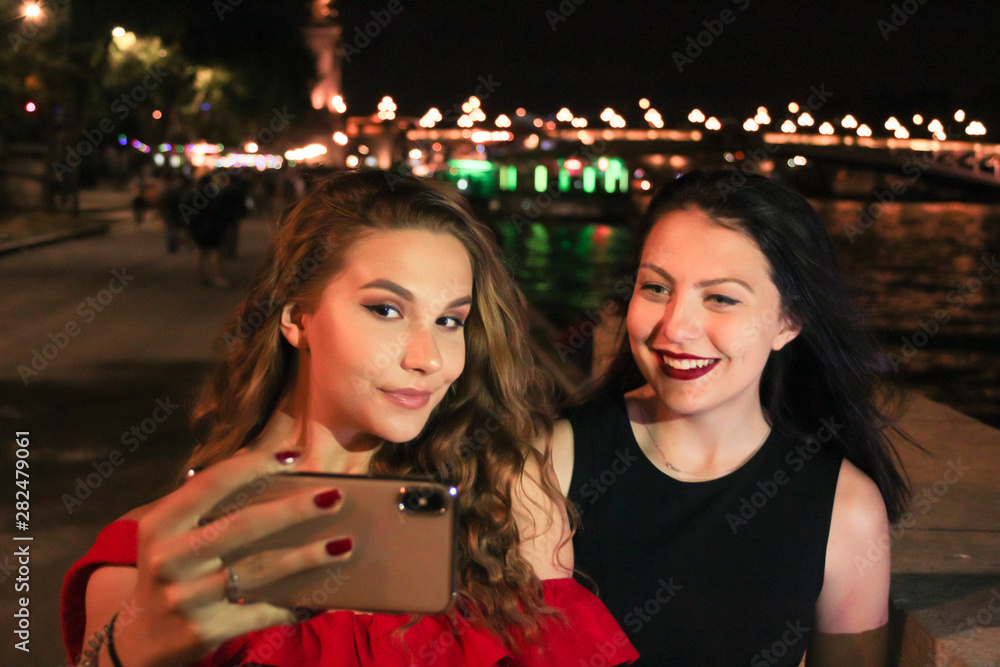 Two pretty girls taking a selfie with a smartphone on vacation. Beautiful blonde woman with red dress and brunette girl in black dress. Happy and smiling. Light of the city in Paris. 