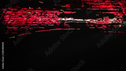 Rainy night. Beautiful red street lighting reflected in rainwater puddle on the road paved wth setts. Rain drops creating beautiful ripples, texture and rhythmical abstract patterns.