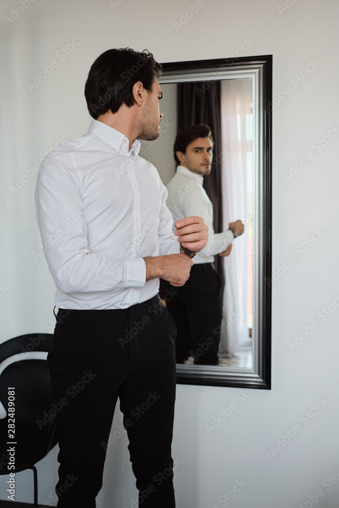 Young handsome brunette man in white shirt and black trousers dreamily looking in mirror at home