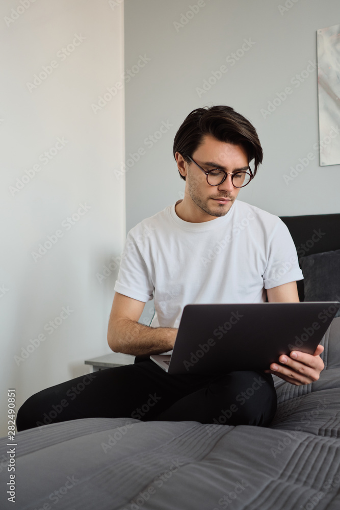 Young serious brunette man in T-shirt and eyeglasses sitting on big bed at modern home thoughtfully using laptop Young serious brunette man in T-shirt and eyeglasses sitting on big bed at modern home thoughtfully using laptop