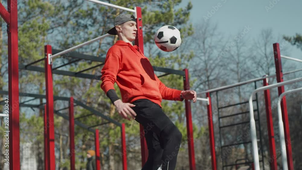 A young guy throws a soccer ball with his knees. Teenager warming up on the background of the Playground, playing football. The boy with the ball near the horizontal bars. Street football. Slow motion A young guy throws a soccer ball with his knees. Teenager warming up on the background of the Playground, playing football. The boy with the ball near the horizontal bars. Street football. Slow motion
