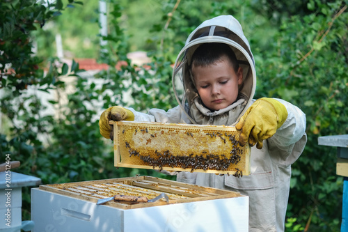 Cheerful boy beekeeper in protective suit near beehive. Honeycomb with honey. Organic food concept. The most useful organic honey. photo