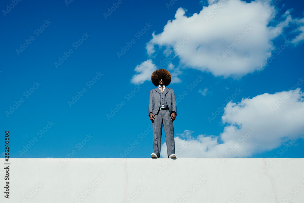 Portrait confident, cool, well-dressed young man with afro standing on wall against sky Portrait confident, cool, well-dressed young man with afro standing on wall against sky
