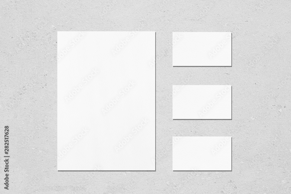 One empty white vertical a4 sized poster and three horizontal rectangle business card mockups with soft shadows on neutral light grey concrete wall background. Flat lay, top view One empty white vertical a4 sized poster and three horizontal rectangle business card mockups with soft shadows on neutral light grey concrete wall background. Flat lay, top view