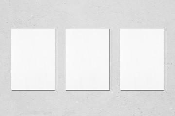 Three empty white vertical rectangle poster mockups with soft shadows on neutral light grey concrete wall background. Flat lay, top view