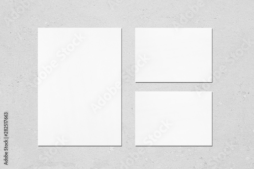 One empty white vertical a4 sized poster and two a5 sized horizontal rectangle card mockups with soft shadows on neutral light grey concrete wall background. Flat lay, top view