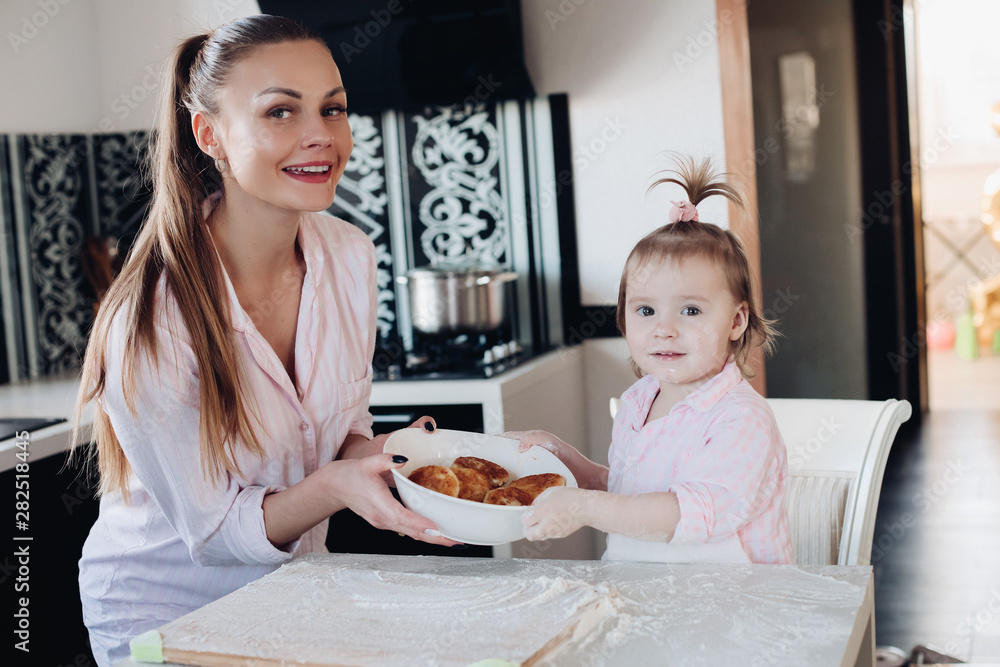 Beautiful woman with lovely child in pajamas holding big bowl with cookies. Close up of happy mother and daughter sitting together at kitchen in morning. Little kid helping parent cooking breakfast.