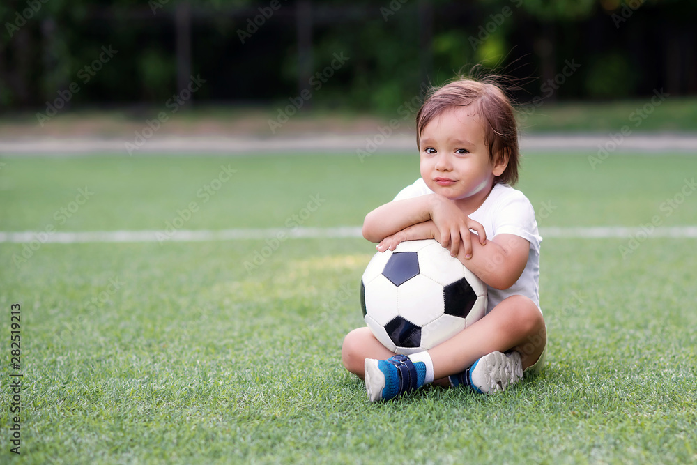 Portrait of sad unhappy boy sitting on football field and holding with both hands or embracing soccer ball. May be his team lost or he wanted to play but was rejected. Competition emotions concept