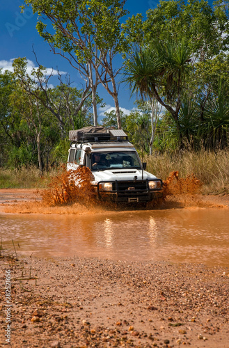 Western Australia – Flooded Outback gravel road with 4WD car crossing the waterhole with splashing muddy water at the savanna