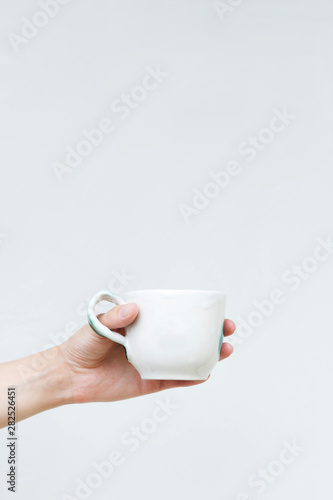 White ceramic mug in a female hand on a background of a white wall. Hand-made