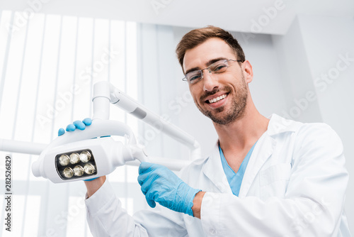 low angle view of cheerful dentist in glasses touching medical lamp and looking at camera photo