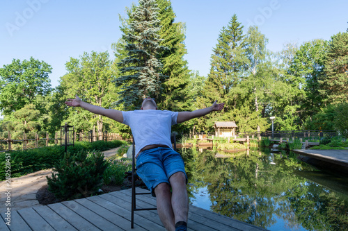 A young man meets the dawn, sitting in front of a pond on a wooden folding chair, spreading his arms wide apart and turning his face to the sky