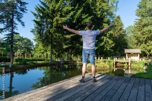A young man stands at dawn in front of a pond with his arms raised to the sky