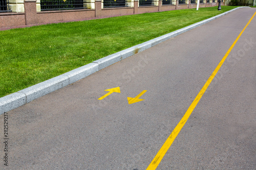 an asphalt road with yellow markings and directional arrows, a roadside with a green lawn and a stone fence.