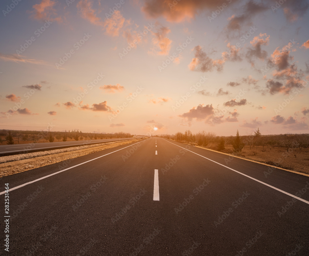 A straight tarmac road at sunset or sunrise. A country motorway stretching out under a dramatic orange sky A straight tarmac road at sunset or sunrise. A country motorway stretching out under a dramatic orange sky