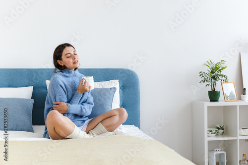 Girl in a blue sweater in interior Hygge style with a cup in hand sits on the bed