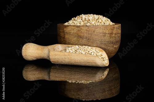 Lot of whole unpeeled sesame seeds in wooden bowl with wooden scoop isolated on black glass photo