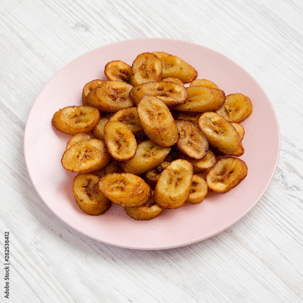 Homemade fried plantains on a pink plate on a white wooden background, side view. Homemade fried plantains on a pink plate on a white wooden background, side view.