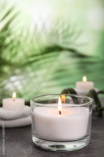 Burning candle in glass holder on grey table against blurred green background, space for text