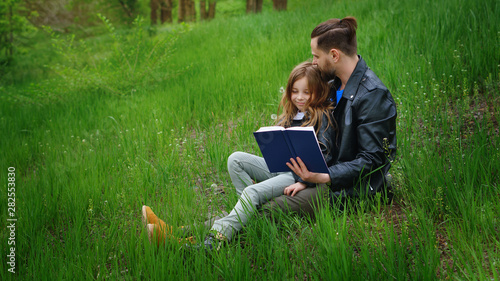 Modern stylish family walking in the park. Father and daughter are reading a book while sitting on a green lawn. Time together. Family look. Urban casual outfit