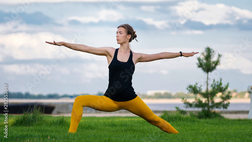Young woman with trim figure practicing yoga warrior pose. Girl performs yoga in a park on International Yoga Day. Yoga helps find balance. Practice asana outdoor. 