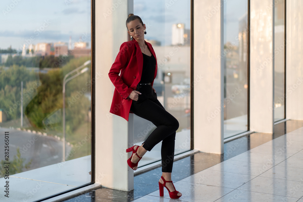 beautiful caucasian girl in red jacket, heels and black trousers stands near panoramic glazing business center. Full height. Office female image. beautiful caucasian girl in red jacket, heels and black trousers stands near panoramic glazing business center. Full height. Office female image.