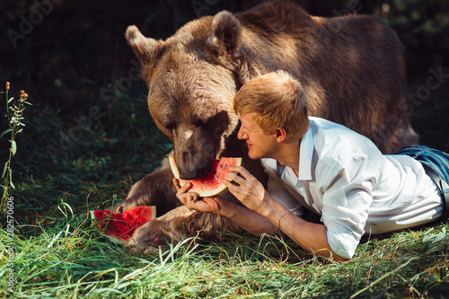 Photography A red-haired man in a white shirt lies next to a brown bear in forest and treats him with a slice of juicy watermelon