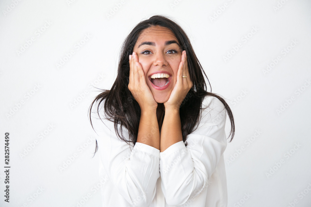 Overjoyed shocked woman rejoicing at awesome news. Happy joyful beautiful young Latin woman in white shirt keeping hands on cheeks, smiling and shouting. Good news or joy concept Overjoyed shocked woman rejoicing at awesome news. Happy joyful beautiful young Latin woman in white shirt keeping hands on cheeks, smiling and shouting. Good news or joy concept