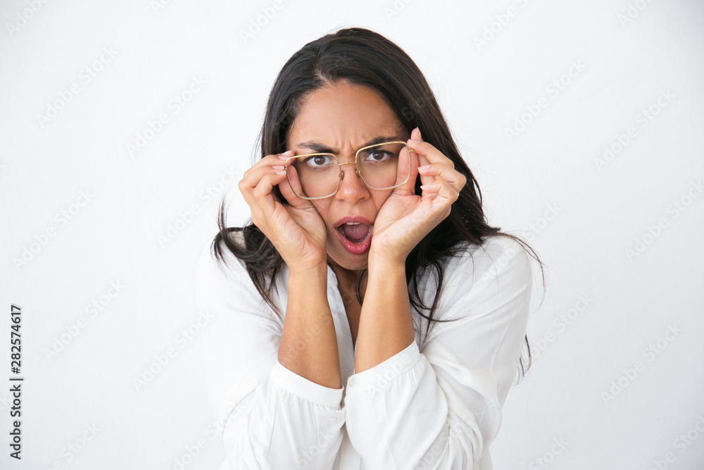 Annoyed exasperated woman putting on eyeglasses. Beautiful young Latin woman in white shirt adjusting glasses and making mad grimace with open mouth. Negative emotions concept Annoyed exasperated woman putting on eyeglasses. Beautiful young Latin woman in white shirt adjusting glasses and making mad grimace with open mouth. Negative emotions concept