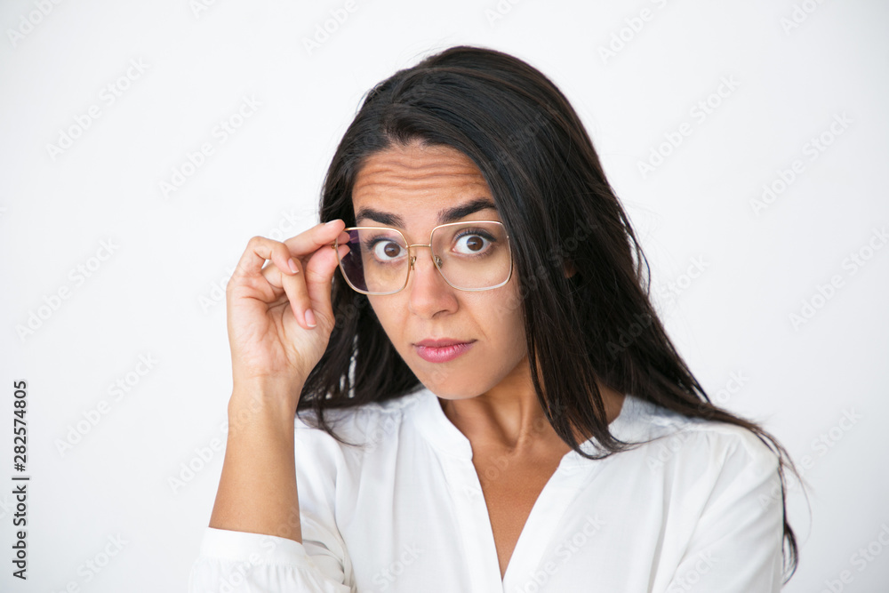 Surprised impressed woman looking at camera through glasses with asking face. Closeup of beautiful young Latin woman in white shirt adjusting eyeglasses. Visionary concept Surprised impressed woman looking at camera through glasses with asking face. Closeup of beautiful young Latin woman in white shirt adjusting eyeglasses. Visionary concept