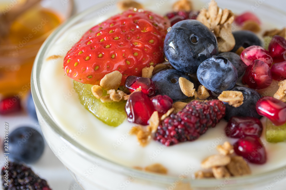 Plain yogurt with strawberry, blueberries, kiwi, granola, pomegranate in a glass bowl and honey on white wooden texture, healthy food and plant-based food concept Plain yogurt with strawberry, blueberries, kiwi, granola, pomegranate in a glass bowl and honey on white wooden texture, healthy food and plant-based food concept