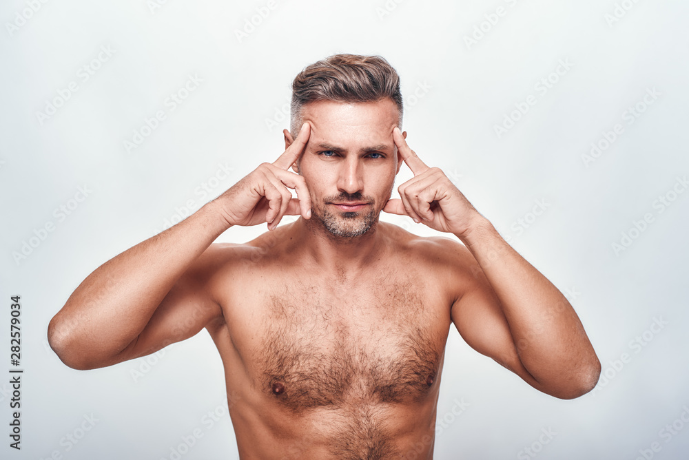 Fototapeta premium Perfection. Handsome and sexy man with a stubble holding head in hands and looking at camera while standing against grey background