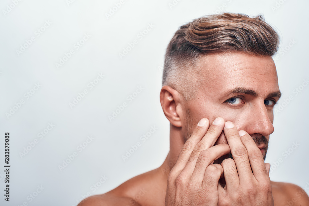 Skin problems. Worried man with a stubble examining his face and looking at camera while standing against grey background Skin problems. Worried man with a stubble examining his face and looking at camera while standing against grey background
