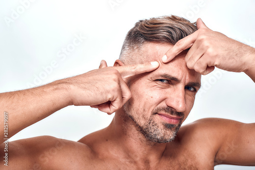 What is wrong with my skin Worried man with a stubble examining his face and looking at camera while standing against grey background