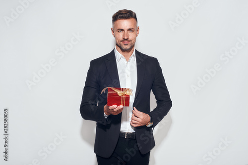 This is for you. Handsome bearded businessman in classic suit holding a gift while standing against grey background