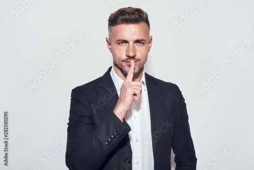 Silence. Portrait of bearded businessman looking at camera and holding finger on lips while standing against grey background