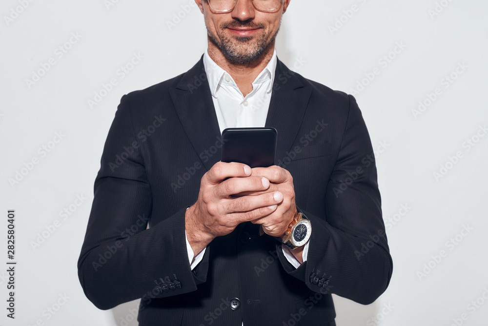 Great news. Portrait of handsome and cheerful bearded man in classic wear and eyeglasses holding smart phone and looking at it while standing against grey background Great news. Portrait of handsome and cheerful bearded man in classic wear and eyeglasses holding smart phone and looking at it while standing against grey background