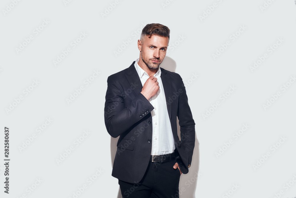 Stylish man. Bearded businessman in classic suit adjusting shirt and looking at camera while standing against grey background Stylish man. Bearded businessman in classic suit adjusting shirt and looking at camera while standing against grey background