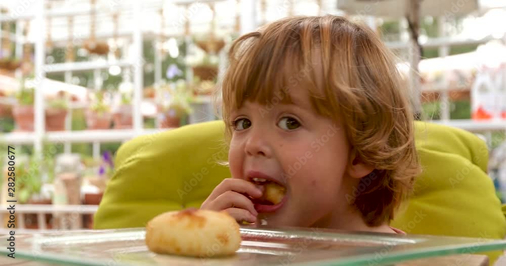 Smart cute kid putting bread to mouth and looking along at table in outside cafe Smart cute kid putting bread to mouth and looking along at table in outside cafe