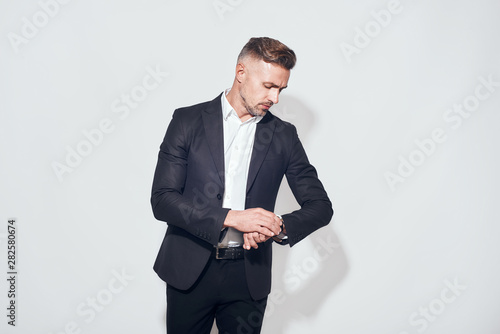 Time to go. Handsome bearded businessman in classic suit looking at watch on his hand while standing against grey background