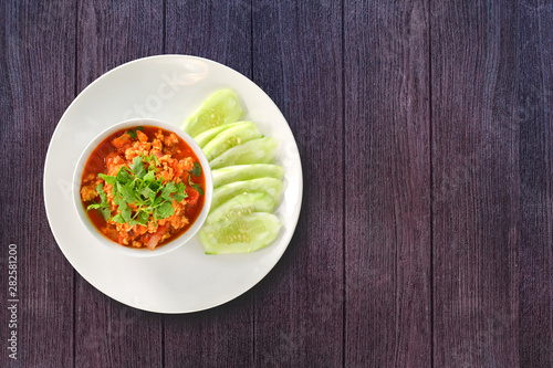 Thai Northern Style Pork and Tomato Relish in white bowl served with cucumber sliced on dark wooden background. Hot and spicy dipping. Top view.