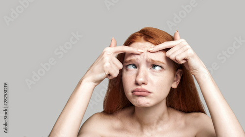 Stop acne. Worried young redhead woman examining her face while standing against grey background photo