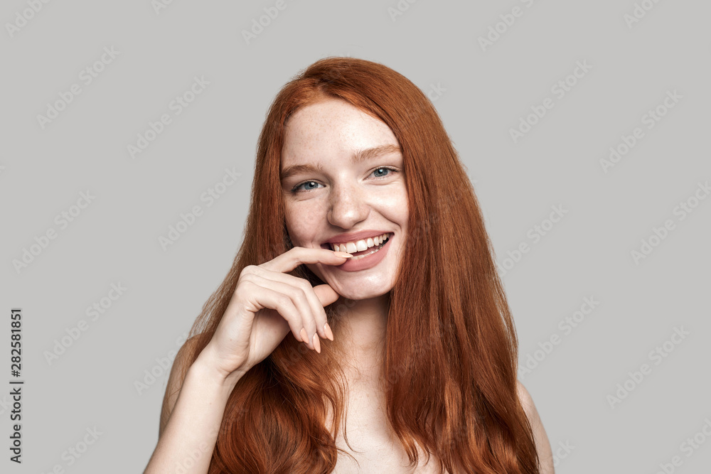 Living with smile. Studio shot of cheerful pretty young redhead lady smiling at the camera while standing against grey background Living with smile. Studio shot of cheerful pretty young redhead lady smiling at the camera while standing against grey background