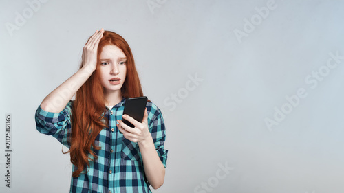 Oh no. Shocked redhead woman in casual wear keeping hand on head and looking at mobile phone while standing against grey background