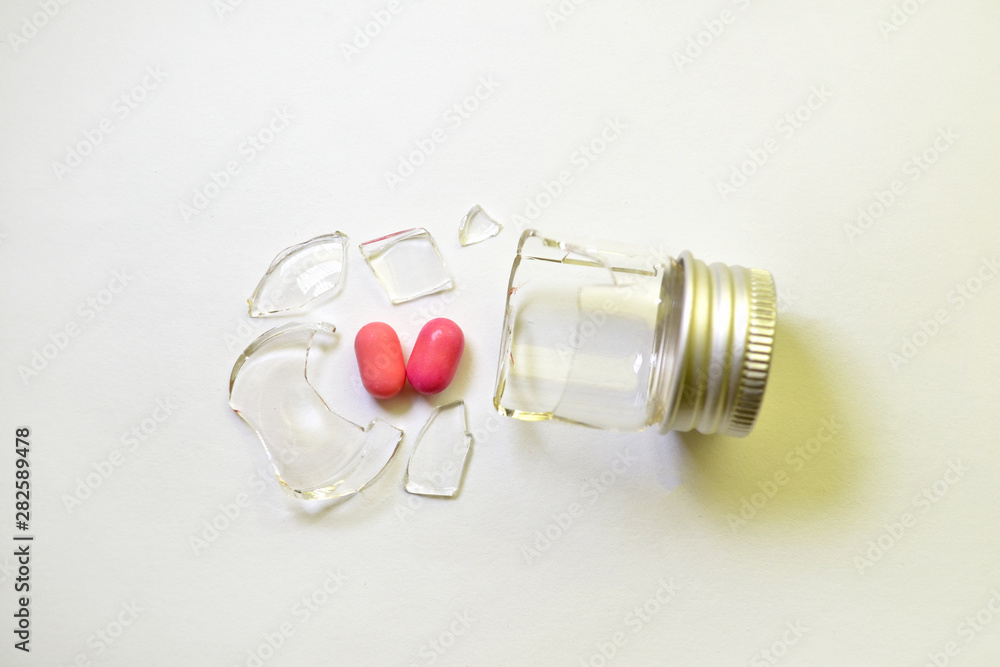 A broken small glass bottle with metal cap, two pink heart-shaped tablets fell out of it, on white background, top view A broken small glass bottle with metal cap, two pink heart-shaped tablets fell out of it, on white background, top view